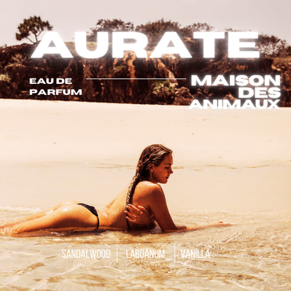 A person lies face down on the sand at the edge of the water, presenting Aurate eau de parfum by Maison des Animaux with scent notes of Sandalwood, Labdanum, and Vanilla.